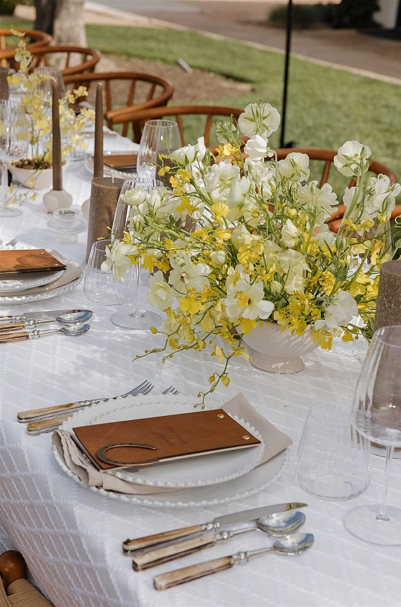 Reception tablescape with an outdoor reception table set in white linens, yellow floral compote, taper candles, glass goblets, and scalloped plates on a lawn