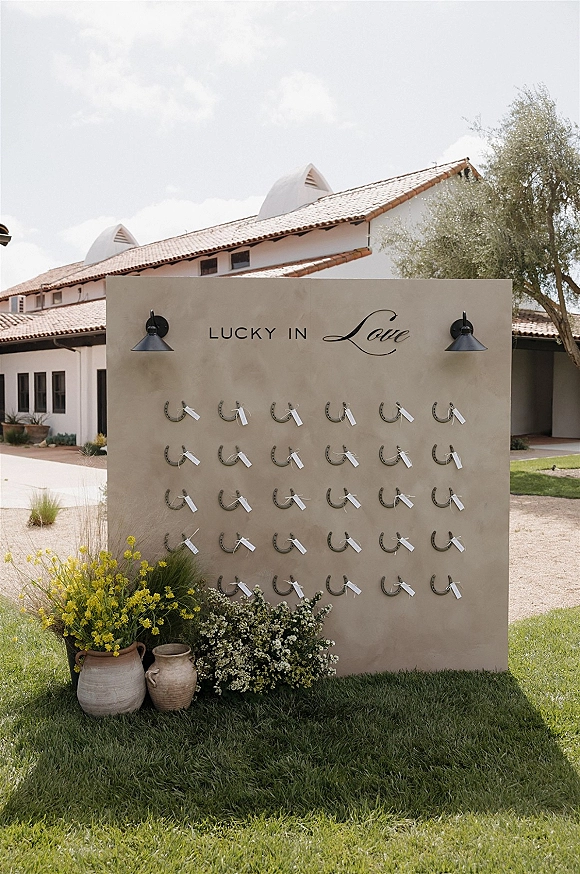Wedding escort display with horseshoe escort cards on a stucco wall, “lucky in love” sign, flanked by yellow flowers in ceramic vases outdoors