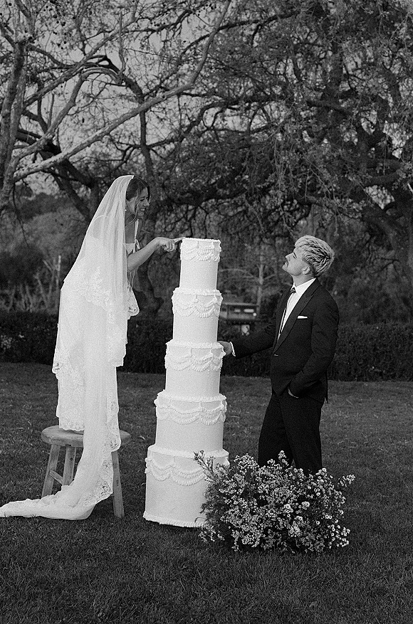 Wedding cake cutting as bride in lace-edged veil stands on a wooden stool to slice a tall tiered cake while groom in tux watches outdoors