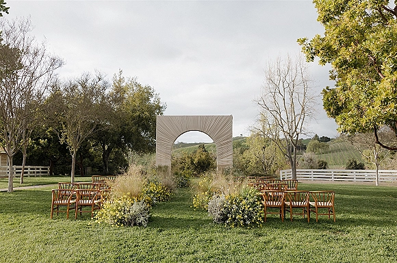 Ceremony setup for an outdoor wedding ceremony with wooden chairs and meadow floral aisle markers leading to a fabric arch on a grassy lawn