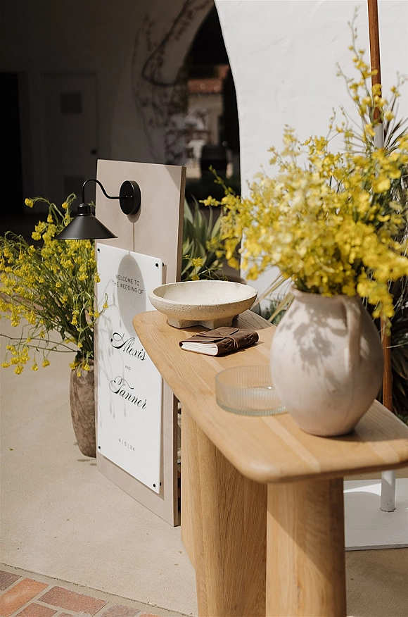 Wedding welcome sign in clear acrylic on a wooden stand beside a guest book on a console table under a stucco archway with yellow florals