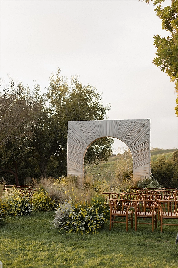 Ceremony setup with draped fabric arch and wooden chairs, lined with meadow florals on a grass lawn overlooking vineyard hills
