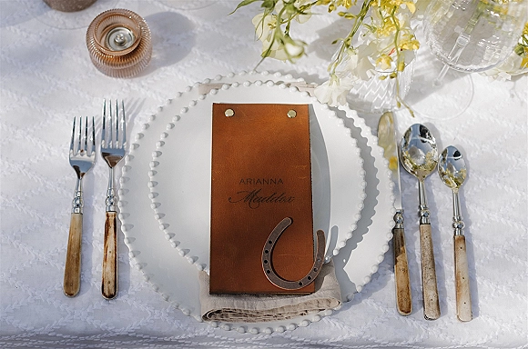 Reception tablescape with wedding place setting on white lace tablecloth, featuring a beaded charger, leather menu, horseshoe, and white-yellow flowers