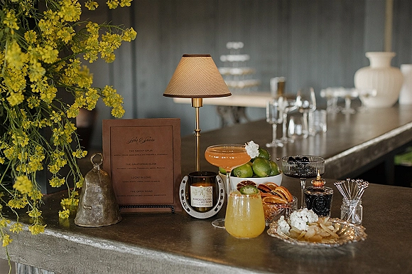 Wedding bar setup with signature cocktail menu, candle, mini lamp, yellow florals, coupe glasses, and lime-and-citrus garnish bowls on a dark bar counter
