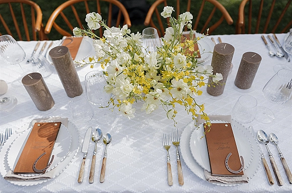 Reception tablescape with white floral centerpiece, yellow blooms, brown pillar candles, beaded plates and menus on a lawn outdoors
