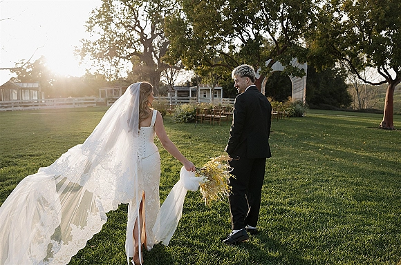 Couple portrait of bride and groom walking hand in hand, bride with long veil and bouquet, backlit on a sunny lawn with trees