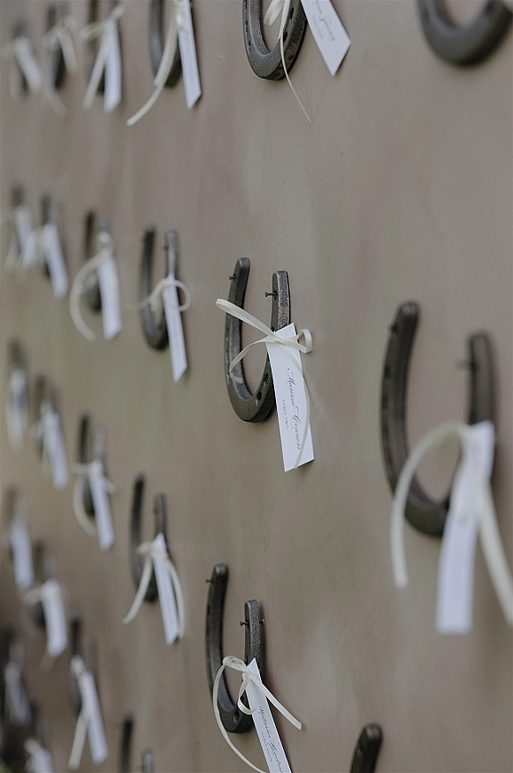 Wedding escort cards with white ribbon bows and name tags hang from horseshoes nailed to a rustic wood board backdrop for seating display