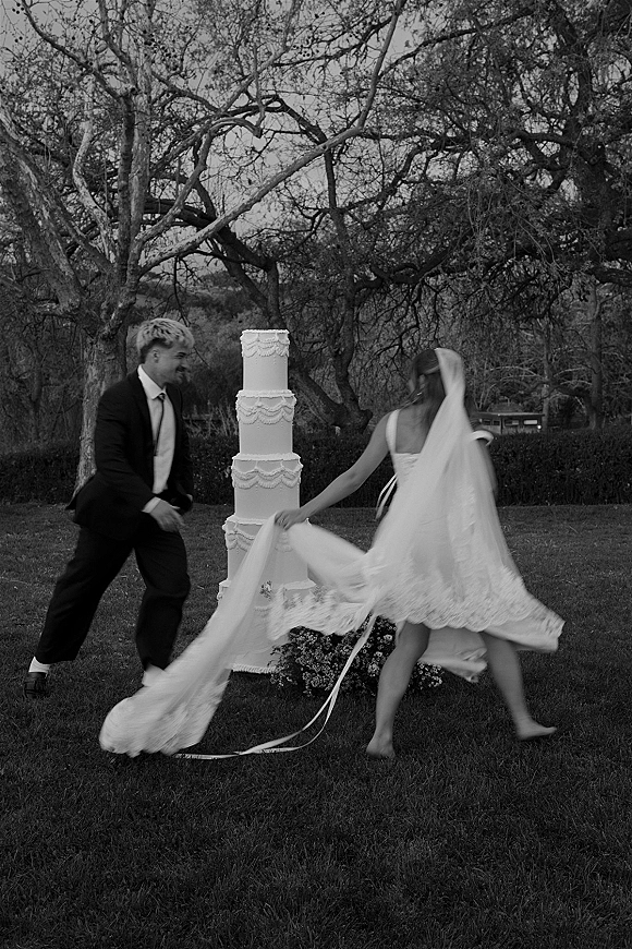 Wedding cake moment as bride in long veil and train plays beside groom in suit near tiered white cake on outdoor lawn with trees