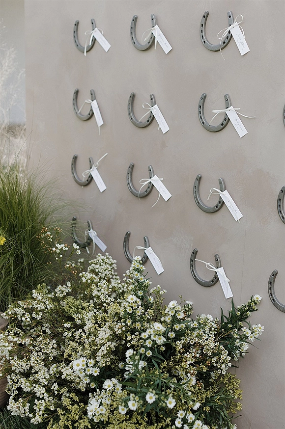 Escort card display with horseshoe escort cards tied in white ribbon, accented by daisies and greenery against a beige wall