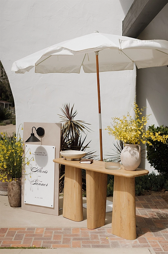 Wedding welcome sign on a pedestal stand beside a wood console table with a ceramic vase of yellow flowers on a sunlit brick patio