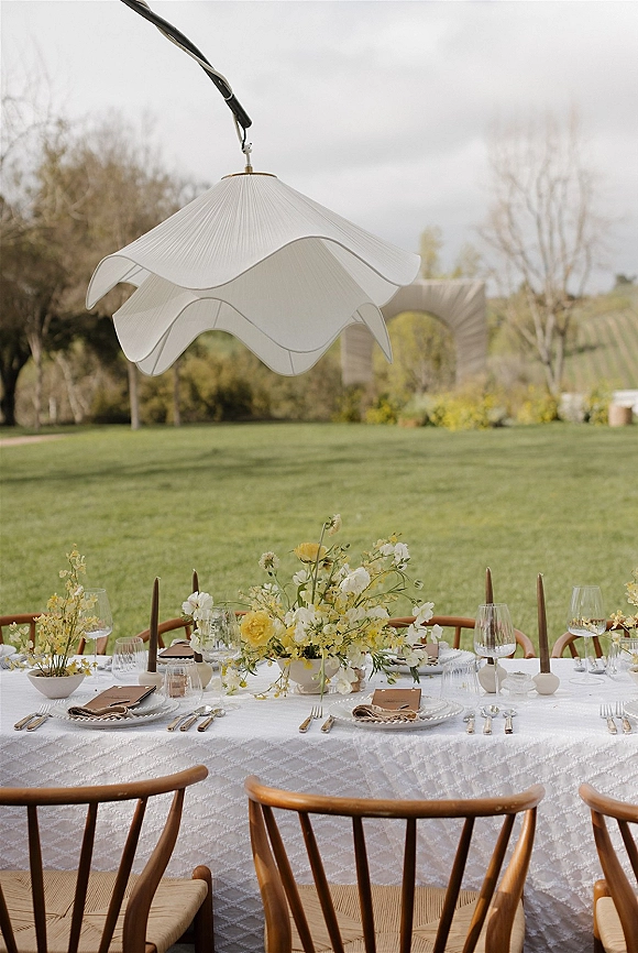 Reception tablescape with an outdoor reception table featuring yellow and white florals, taper candles, and place settings on a cloudy lawn