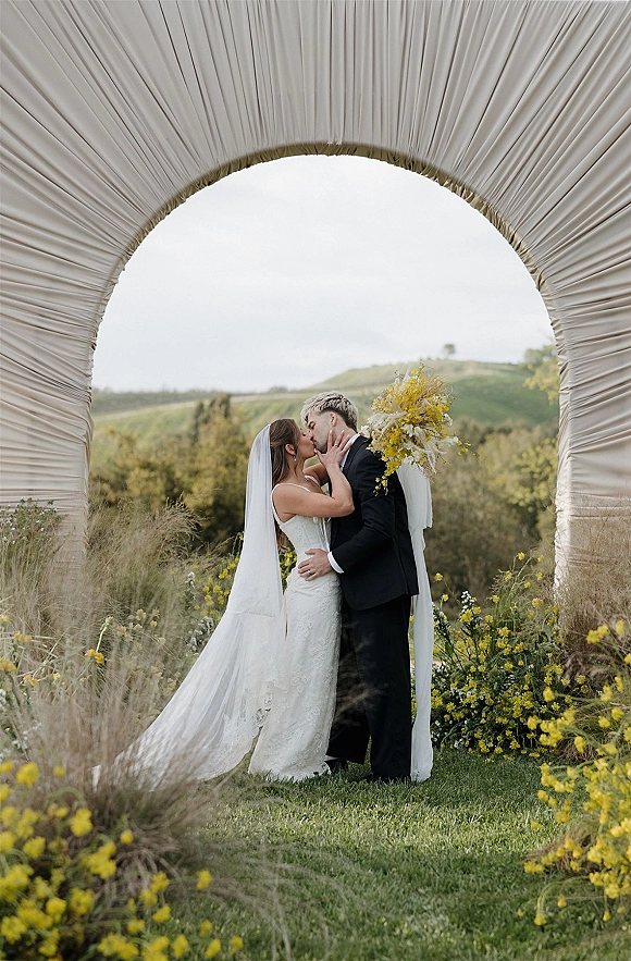 Wedding kiss portrait of bride and groom kiss beneath a floral arch with draped fabric, her long veil and yellow bouquet in a wildflower meadow