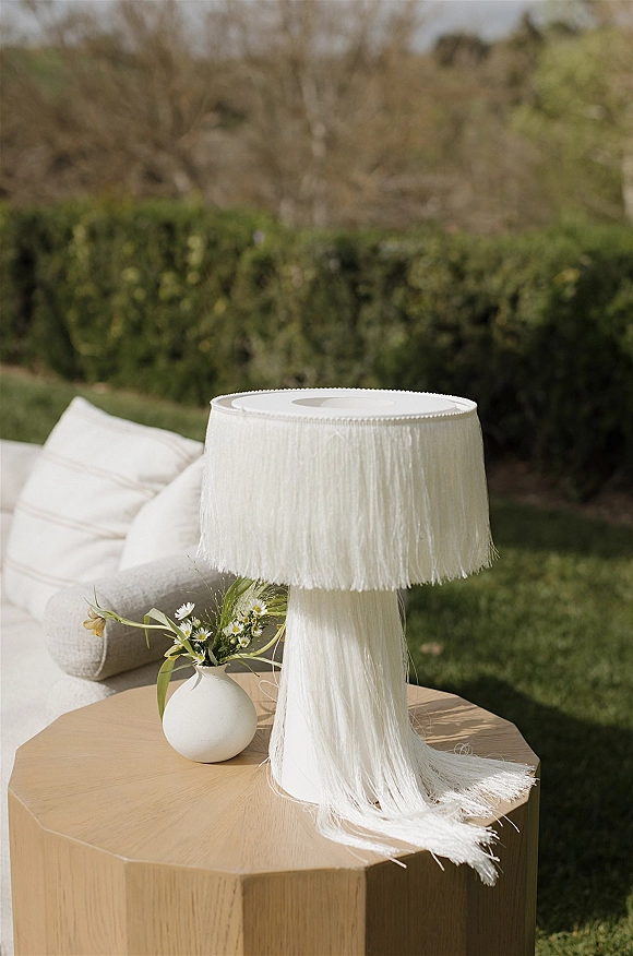 Wedding lounge decor with a white outdoor sofa, throw pillows, and fringe lamp on a wooden side table with bud vase flowers on a lawn