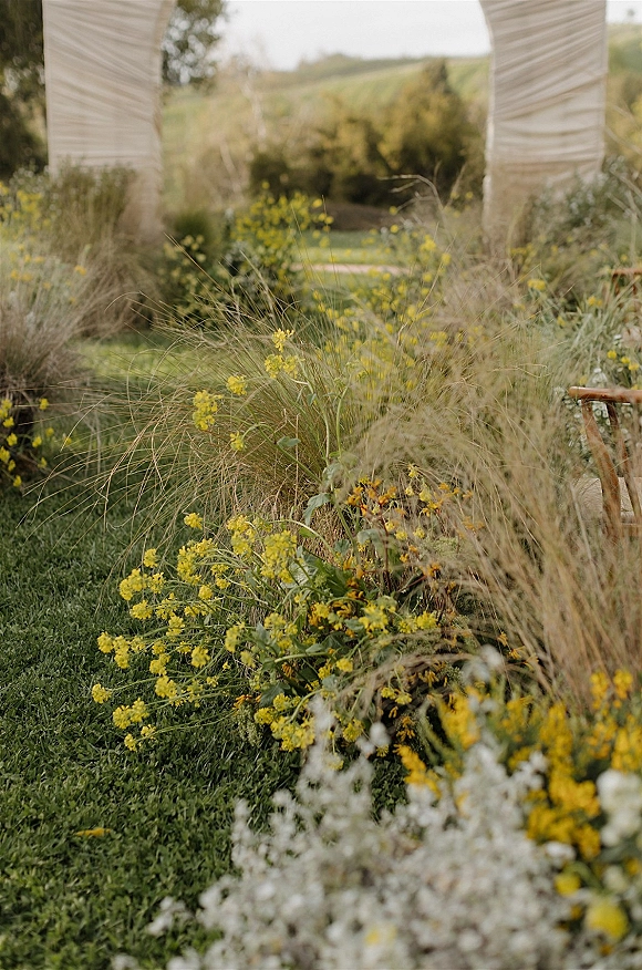 Ceremony aisle decor with meadow aisle flowers, wildflowers and meadow grass leading to a white draped fabric arch on a hillside lawn