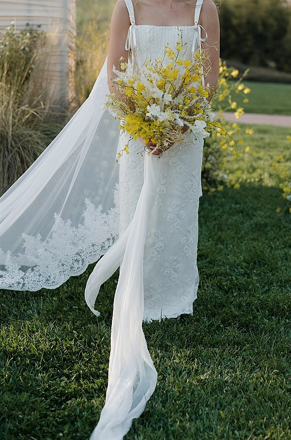 Bridal bouquet of yellow wildflowers with white blooms and greenery held against a white lace dress and veil on a garden lawn path