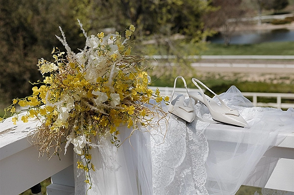 Bridal shoes, white slingback heels with bows beside a lace veil and yellow-white bouquet on a railing overlooking greenery and pond