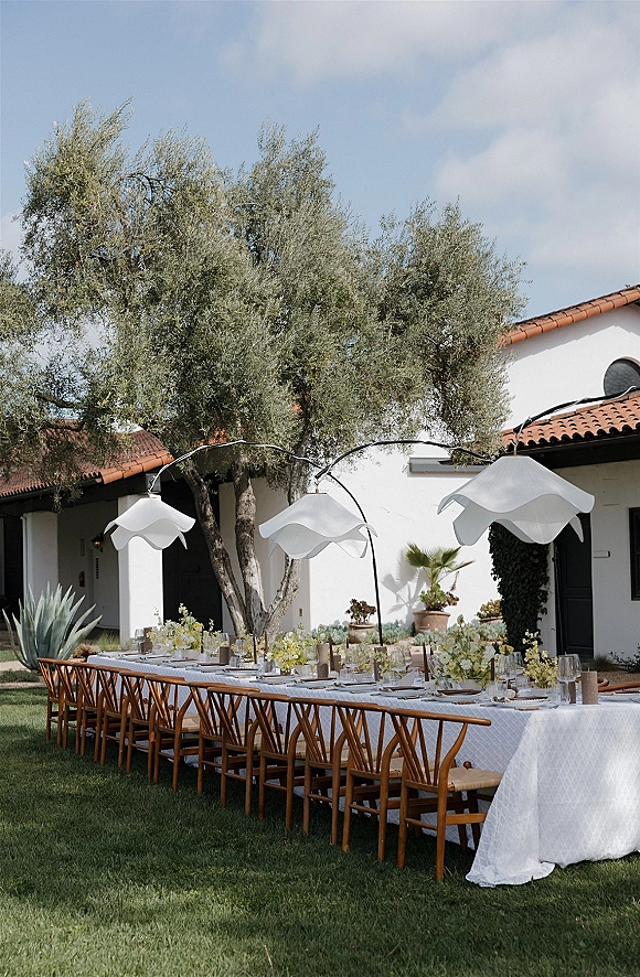 Reception tablescape for an outdoor reception tablescape with white tablecloth, wooden chairs, florals, bud vases, and candles under sculptural lights.