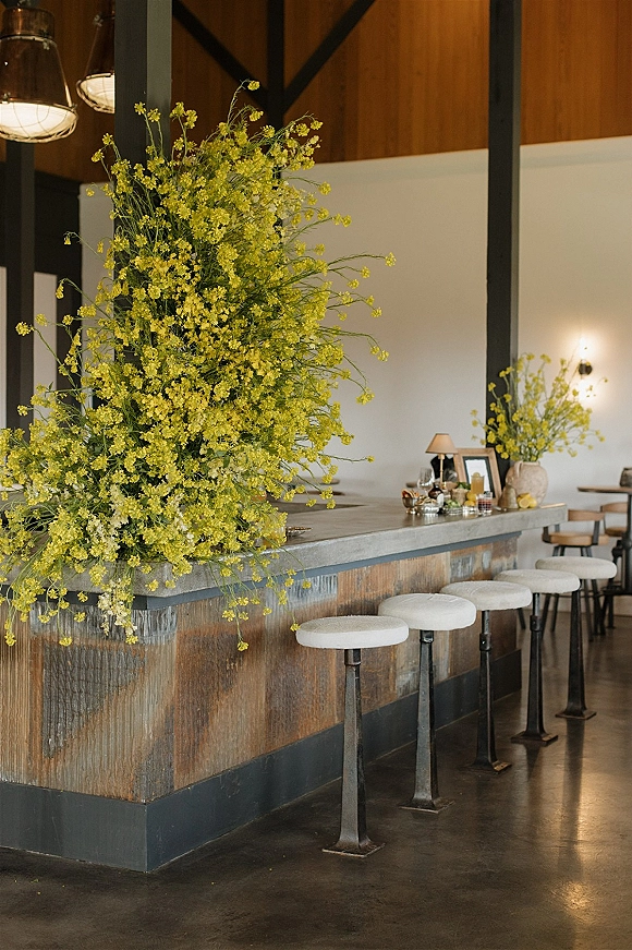 Reception bar decor with a yellow floral installation above a styled counter of candles, framed photos, and glassware on a wood-paneled wall background