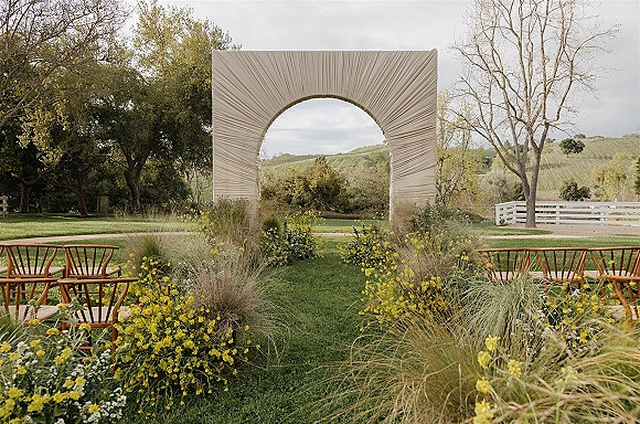 Ceremony setup for an outdoor wedding ceremony with a fabric-draped arch, wooden chairs, and yellow aisle florals on a grassy lawn by a white fence