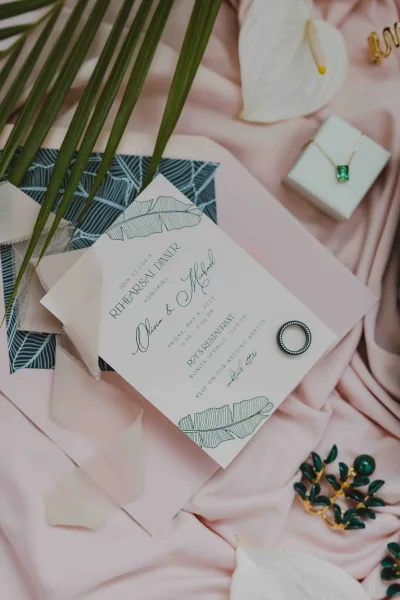 Wedding invitations arranged as a wedding invitation suite on pink satin, with vellum wrap, palm leaf, anthurium flowers, and ring accents
