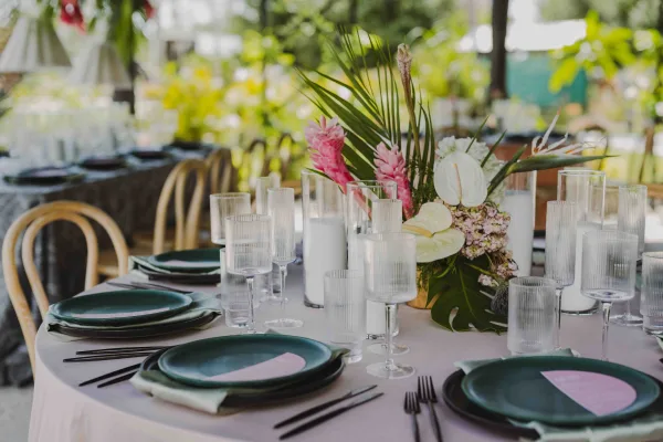 Reception tablescape with tropical wedding tablescape centerpiece of palm leaves, anthurium and pink ginger on an outdoor table with greenery behind