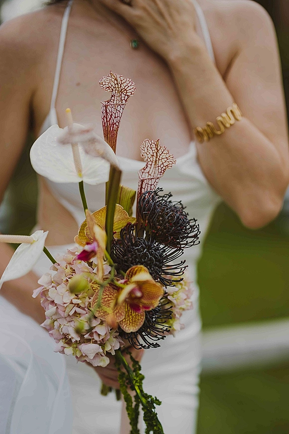 Bridal bouquet with tropical bridal bouquet blooms—anthurium, orchids, hydrangea, and dark protea—held against a white dress in outdoor greenery