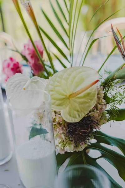 Reception centerpiece with tropical wedding centerpiece blooms—white anthurium, hydrangea, protea, and palm fronds in a glass cylinder vase with candle on white linen