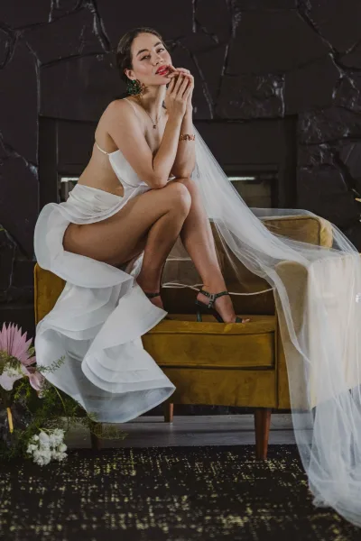 Bridal portrait of a bride in a white slip wedding dress with a long veil, seated on a velvet chair against a dark textured wall