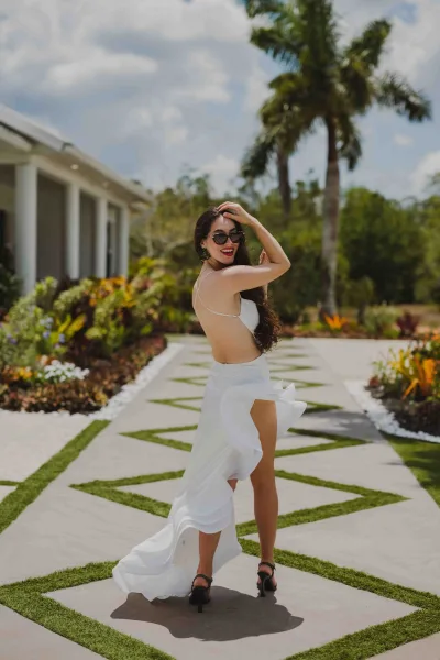 Bridal portrait of a bride in sunglasses wearing an open-back white gown with a thigh-high slit, walking a palm-lined garden walkway under blue sky