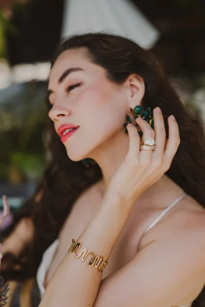 Bridal jewelry close-up with emerald wedding earrings as the bride touches them, showing ring stack and gold bracelet against soft greenery