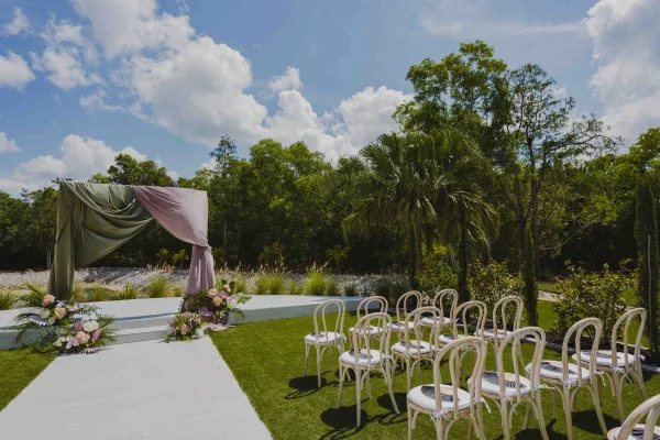 Ceremony setup for an outdoor wedding ceremony with draped fabric arch, pastel florals, and rows of chairs on a sunny lawn with palms