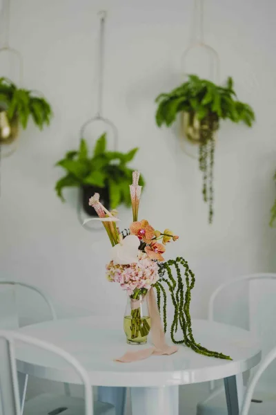 Wedding centerpiece with orchids and hydrangeas in a clear glass vase, tied with blush ribbon on a round white table with hanging greenery