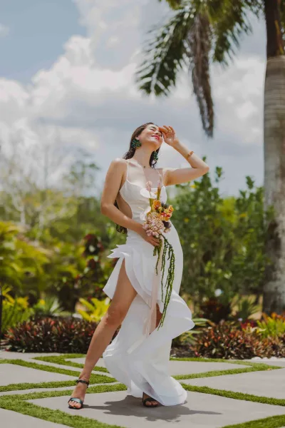 Bridal portrait of a bride holding anthurium bouquet in a strapless white dress with high slit, walking a tropical garden path under blue sky