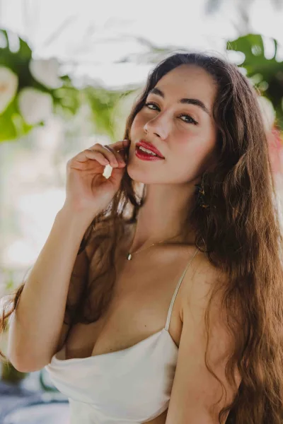 Bridal portrait of a bride with red lip bridal makeup in a white satin slip dress, gold necklace and drop earrings against blurred greenery