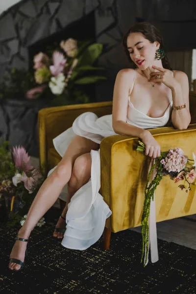 Bridal portrait of a bride on a velvet sofa in a white cowl-neck dress, holding a bouquet with pink ribbon against a moody dark wall