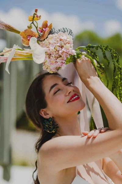 Bridal portrait of a bride holding bouquet with pink hydrangea and orchids, eyes closed and red lipstick against sky and greenery backdrop