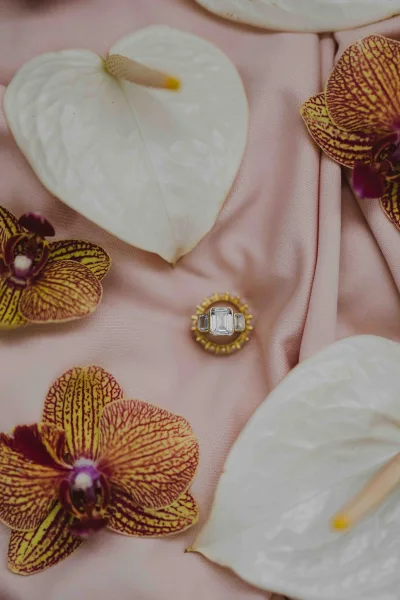 Engagement ring with an emerald cut engagement ring in a gold setting nestled among orchids and anthurium on a pink fabric backdrop