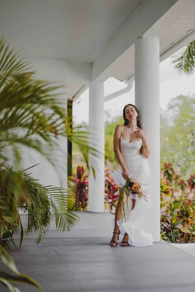 Bridal portrait of a bride leaning on a white column, holding a bouquet in a white slip dress with statement earrings and black heels on a tropical porch