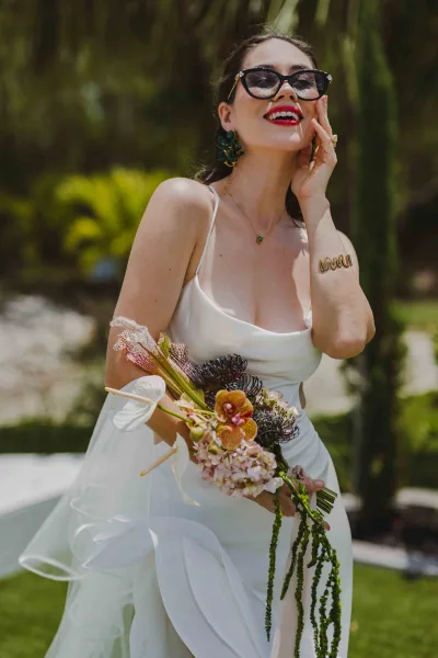Bridal portrait of a bride wearing sunglasses in a white slip wedding dress and veil holding a bouquet in a sunlit garden setting