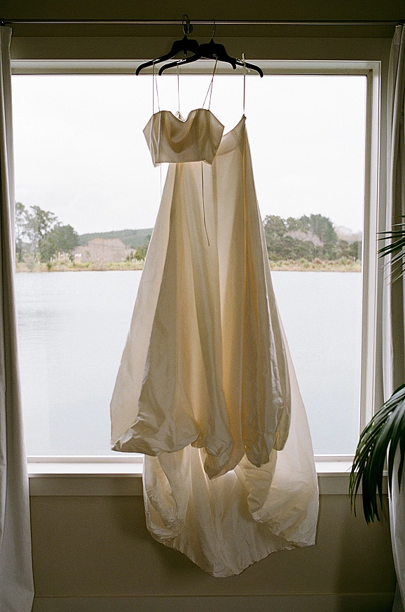 Wedding dress in satin hanging on a dress hanger by a window, with curtains framing a lake and distant trees beyond the shore