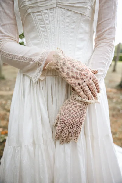 Bridal gloves in white crochet with lace cuffs on a long sleeve corset wedding dress, shown in soft natural light on a leafy path