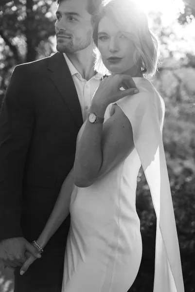 Couple portrait in a black and white wedding portrait style, bride holding groom’s hand with cape sleeve gown in sunlit trees backdrop