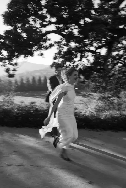 Wedding couple portrait in black and white as bride in sheath gown and groom in dark suit run hand in hand under sunlit tree canopy