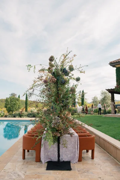Reception tablescape with a tall floral centerpiece and greenery on a patterned runner, set poolside on a stone patio with vineyard hills beyond