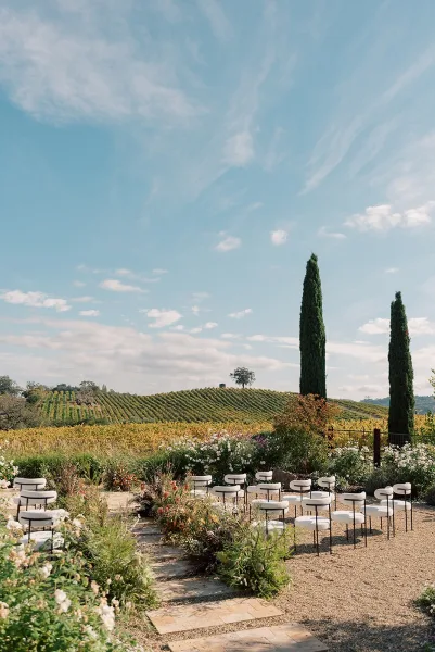 Outdoor ceremony setup with vineyard wedding ceremony chair rows flanking a gravel and stone paver aisle with meadow florals, cypress hills beyond