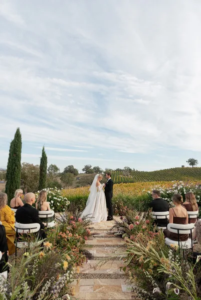 Ceremony moment at an outdoor wedding ceremony as bride and groom exchange vows on a stone aisle, vineyard rows and hills behind