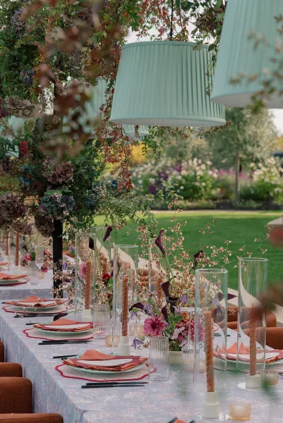 Reception tablescape with an outdoor reception table set with floral centerpiece, spiral taper candles, coral napkins, and lantern shades on a garden lawn