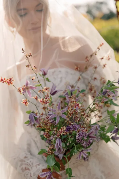 Bridal portrait of a bride holding bouquet with a soft wedding veil over her face, lace dress, and sunlit greenery behind her