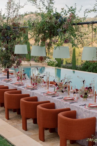 Reception tablescape with patterned tablecloth, floral centerpieces, candles and hanging greenery over a long banquet table beside a pool on a stone patio