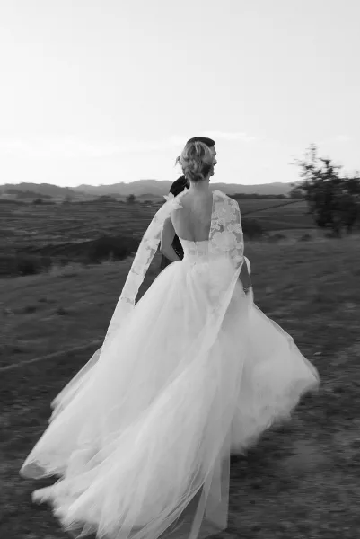 Wedding couple portrait in black and white, bride spinning her flowing tulle gown with lace sleeves beside groom in dark suit and hat in a field